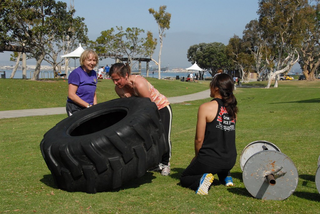 Three women outdoors raising a large tractor tire, with one woman leaning over its elevated edge