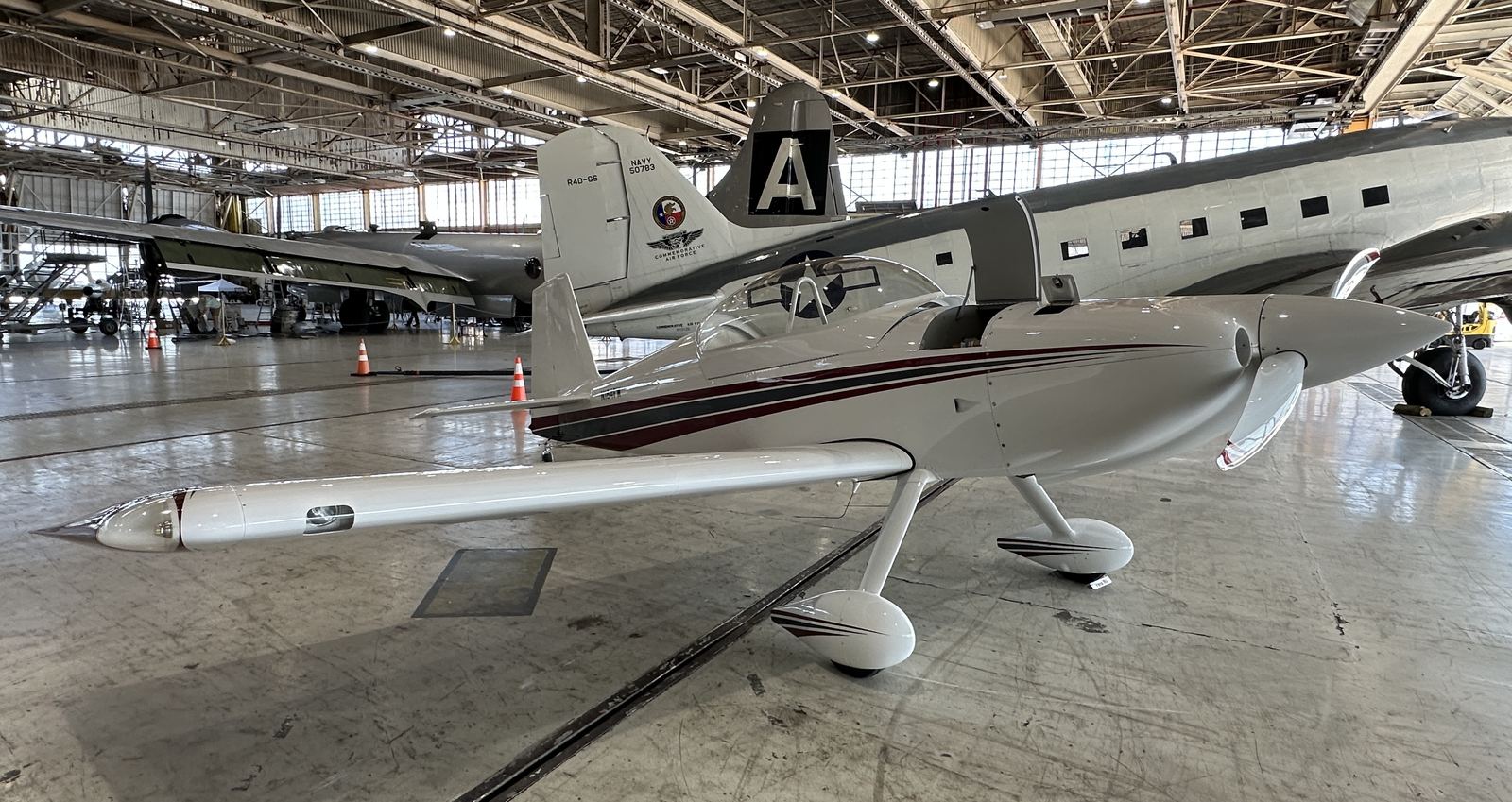 A white single-engine light aircraft parked in a hangar, with a larger military transport aircraft and several people behind it.